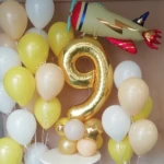 Neunter Geburtstag Dekoration für einen Jungen mit einer goldenen gasgefüllten 9 und einem fliegenden Flugzeug-Folienballon und mehreren gasgefüllten Latex-Ballons in gelb, cremefarben und weiß Latex-Ballons in gelb, creme und weiß mit einer gasgefüllten Folien-Zahl 9 zum Geburtstag und einem Gas gefüllten Figurenballon aus Folien als Flugzeug dargestellt